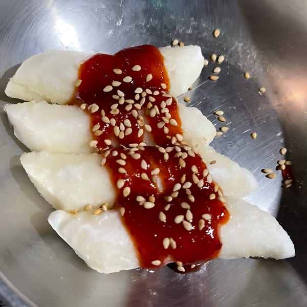 White Korean rice cakes topped with red sauce and sesame seeds in a metal bowl.