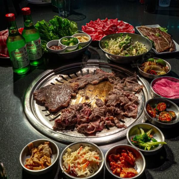 Korean BBQ grill cooking various meats, surrounded by banchan, fresh lettuce, and two bottles of soju.