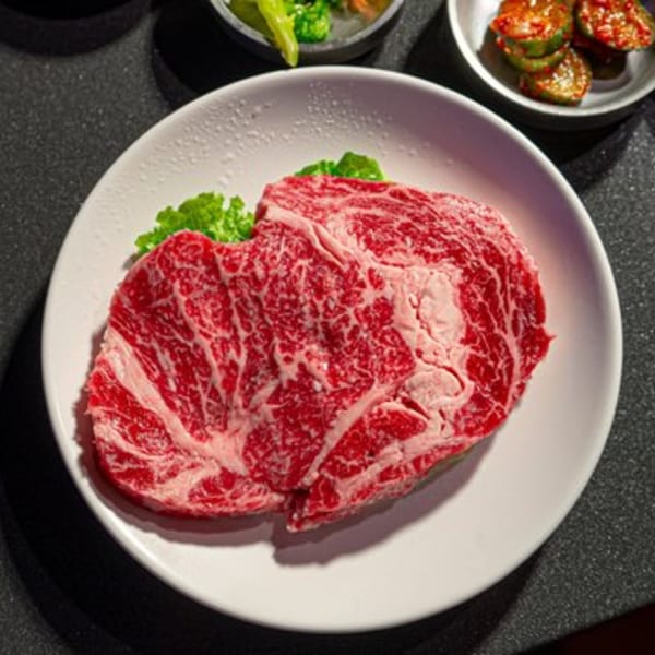 Raw, marbled Korean BBQ beef on a white plate with lettuce, next to side dishes.