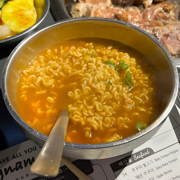 A metal bowl of Korean ramen with green onions, next to a menu and grilled meat.
