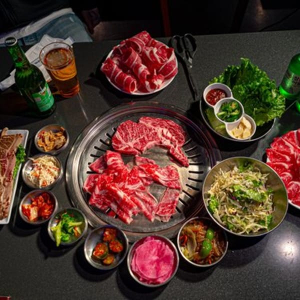 Korean BBQ table with raw meats, banchan, fresh lettuce, sauces, and beverages.