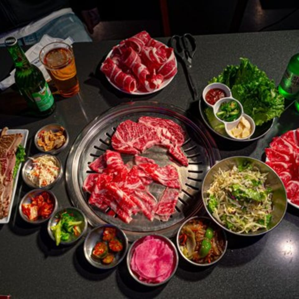 Korean BBQ table with raw meats, banchan, fresh lettuce, sauces, and beverages.